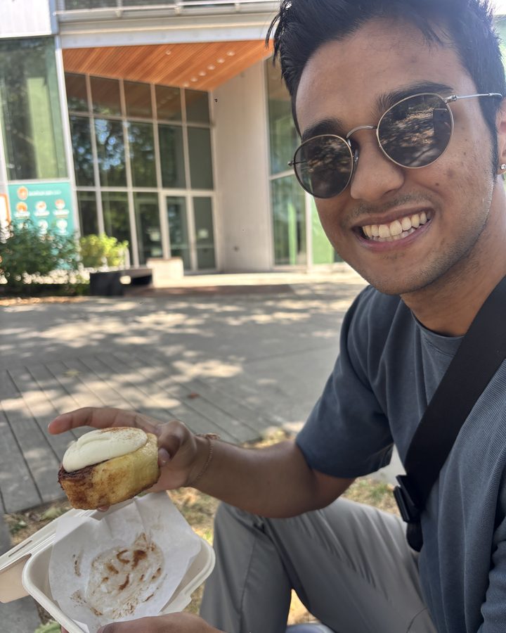 Khushwin outside on a sunny day, smiling, holding a cinnamon bun